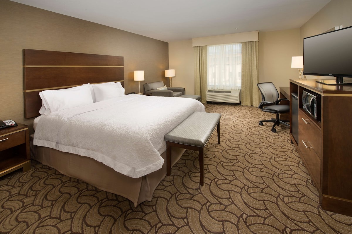 A hotel room with a large bed, two bedside tables, a desk with a chair, a television, and a window with curtains.