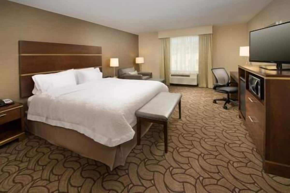 A hotel room with a large bed, bedside tables, a desk, a chair, a television, and a window with curtains.