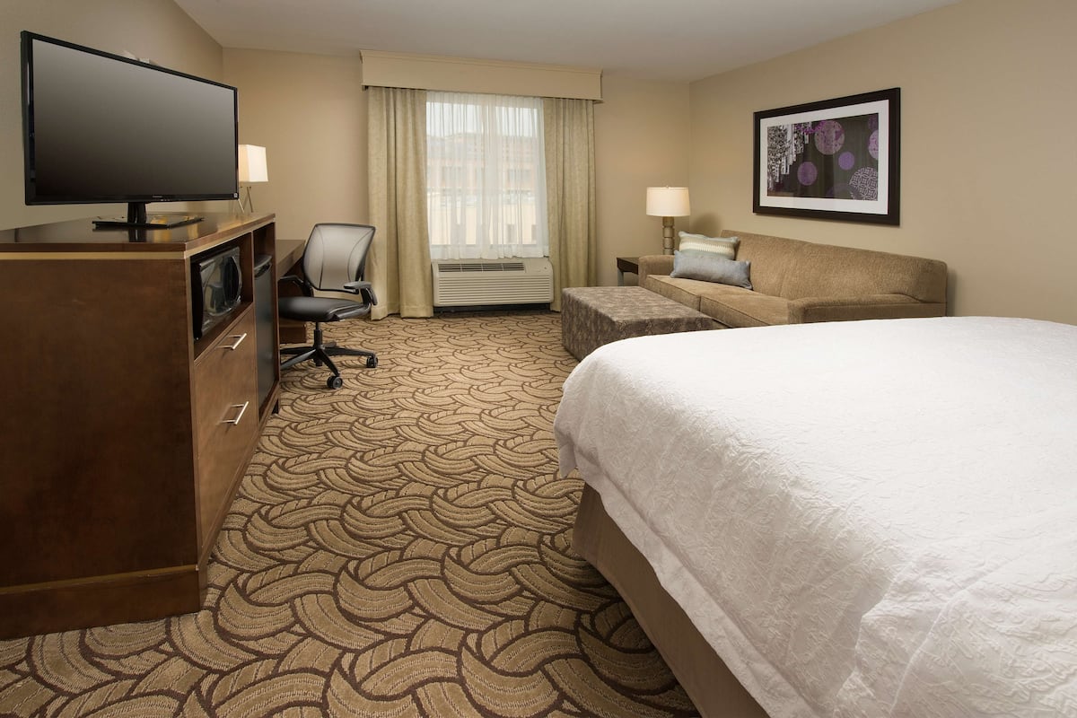 A hotel room with a large bed, a desk with a flat-screen TV, a chair, a sofa, and a framed picture on the wall.