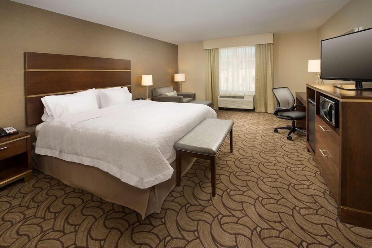 Quarto de hotel com uma cama grande, uma escrivaninha com televisão, uma cadeira e uma luminária de mesa.