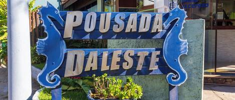 Une enseigne portant le nom « Pousada Daleste », écrite dans une police décorative, est entourée de verdure et d’un jardinier avec des plantes.