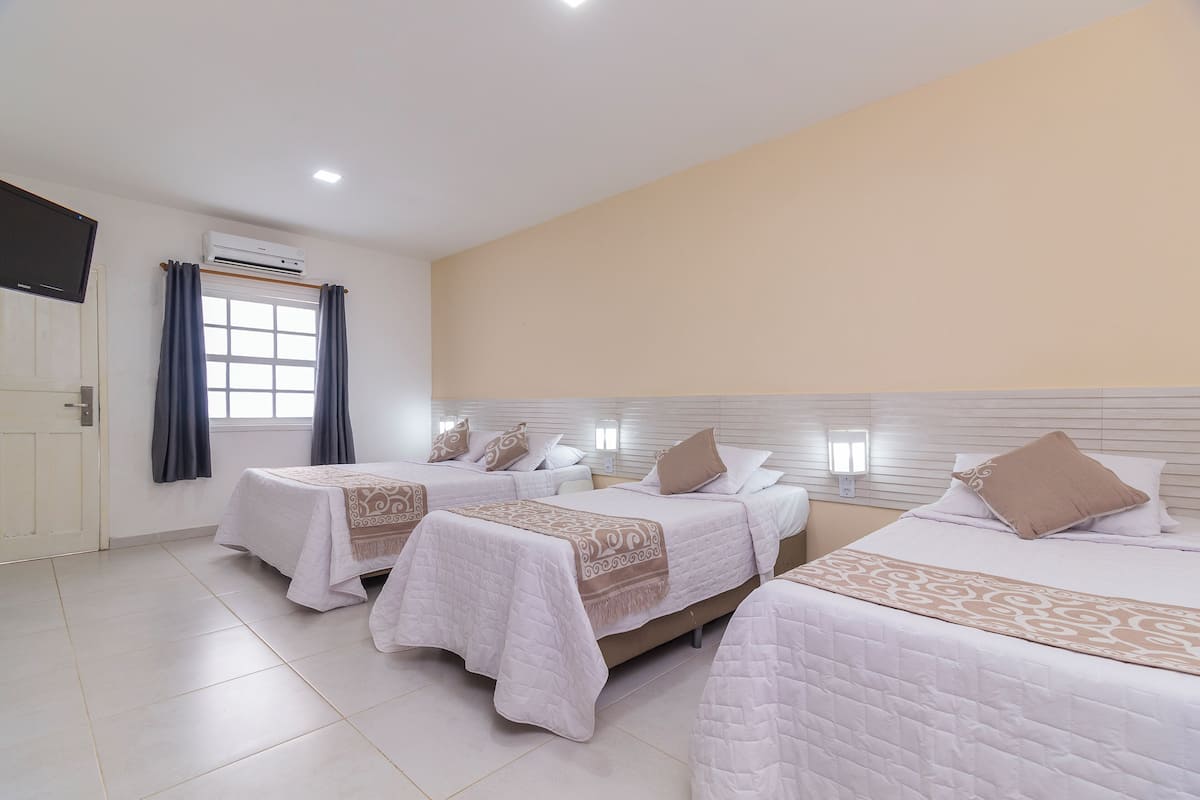 Una habitación de hotel con tres camas, televisión, aire acondicionado y una ventana con cortinas.