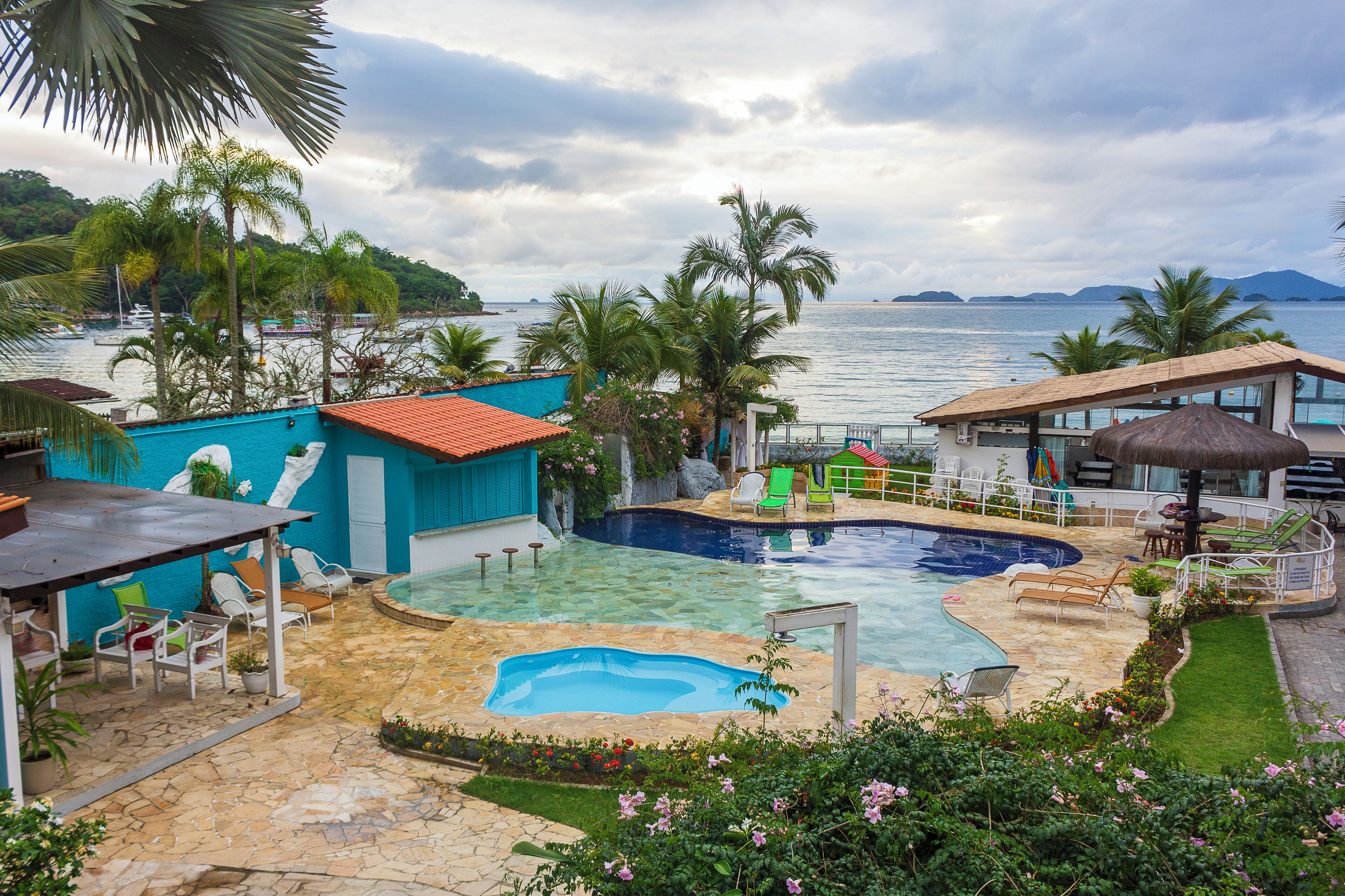 Zona de piscina con vista al mar, rodeada de palmeras y un edificio con techo rojo.