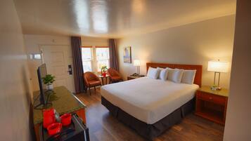 A hotel room with a large bed, a desk with a computer and red mugs, a chair, a nightstand with a lamp, and a window with curtains.