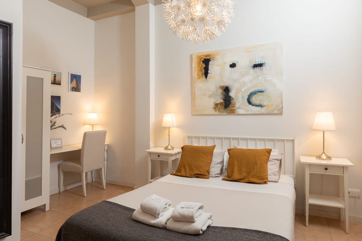 Un dormitorio de hotel con cama, dos mesitas de noche con lámparas, un escritorio con silla, un armario y un cuadro abstracto grande en la pared.