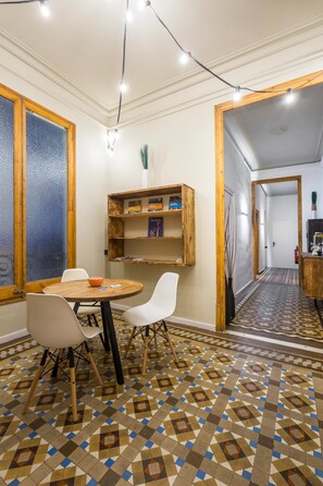 A dining area with a round table, white chairs, a patterned floor, wooden shelves, and a view into another room. - Casa Maca Guest House (Barcelona)