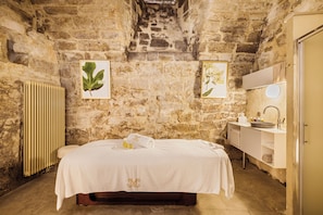 A stone-walled room with a bed, a radiator, a sink, and framed botanical prints.