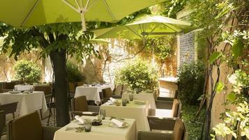 Un espacio al aire libre para comer con mesas y sillas, cubierto por sombrillas verdes, rodeado de vegetación y muros de piedra.