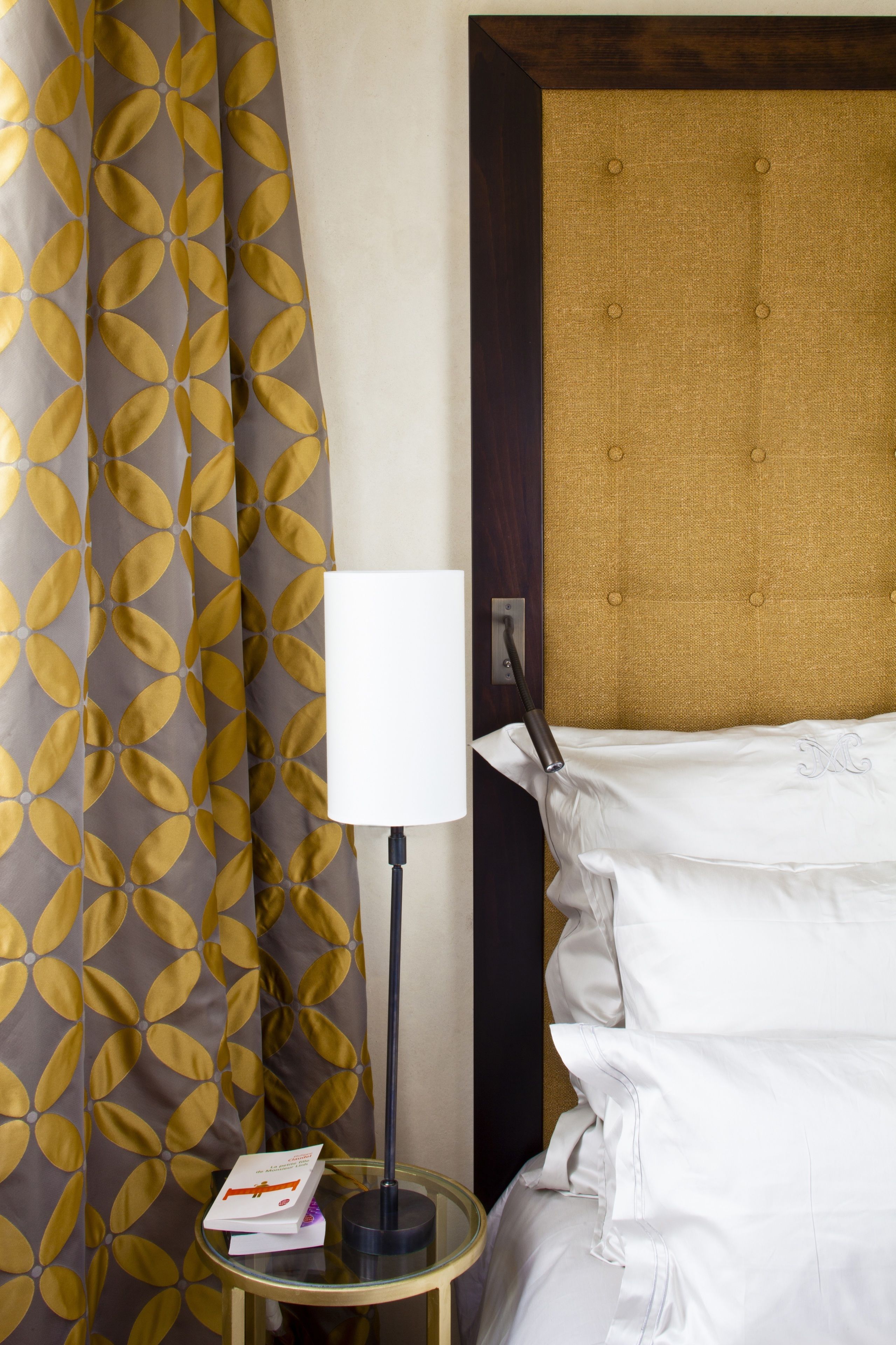 Ein Hotelzimmer mit einem Bett, einer Nachttischlampe, einem kleinen Tisch mit einem Buch und einem Vorhang mit Blätermotiv.