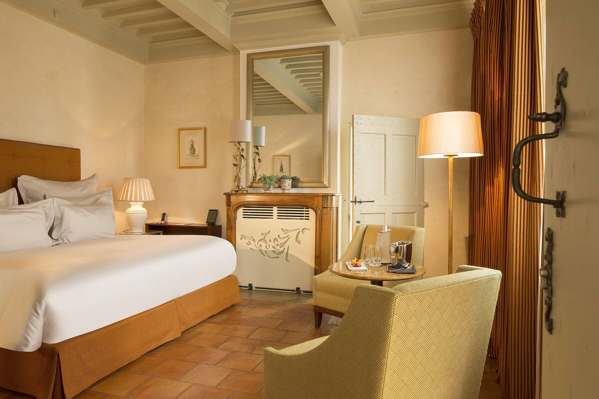 A hotel room with a bed, a chair, a table, a lamp, a mirror, and a door.