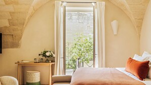 A bedroom with a bed, a small table, a chair, and a window with a view of greenery. - La Maison d'Uzes (Uzès)