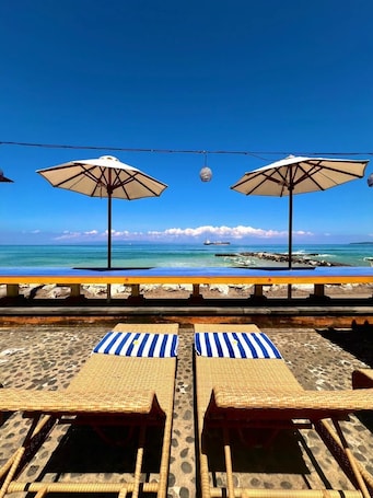 A beachside lounge with two umbrellas, a pool, and lounge chairs.. Ashyana Candidasa