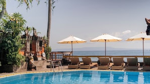 A swimming pool with lounge chairs and umbrellas, surrounded by tropical plants and a view of the ocean. - Ashyana Candidasa (Karangasem)