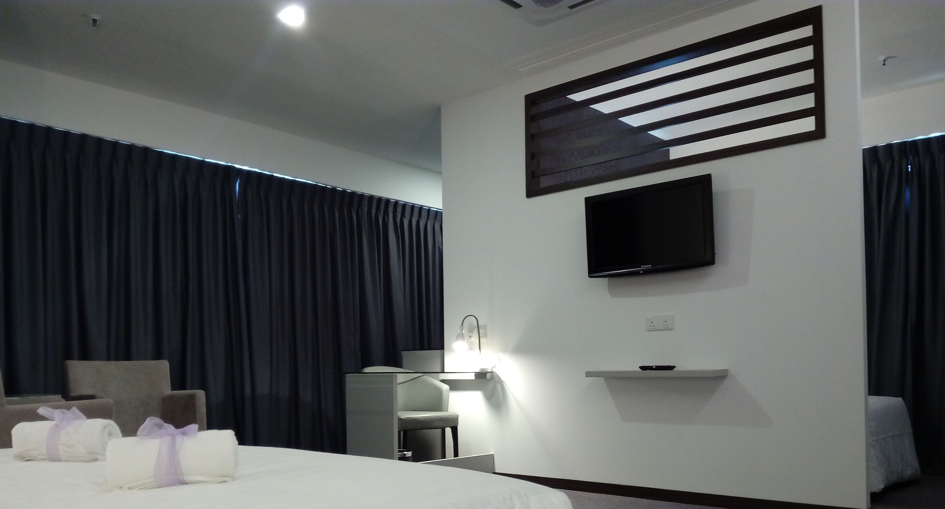 A modern hotel room with a bed, a desk, a chair, a flat-screen TV, and a wall-mounted shelf.