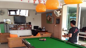 Uma mesa de bilhar com superfície de feltro verde, uma pessoa ao lado da mesa e uma televisão presa na parede.