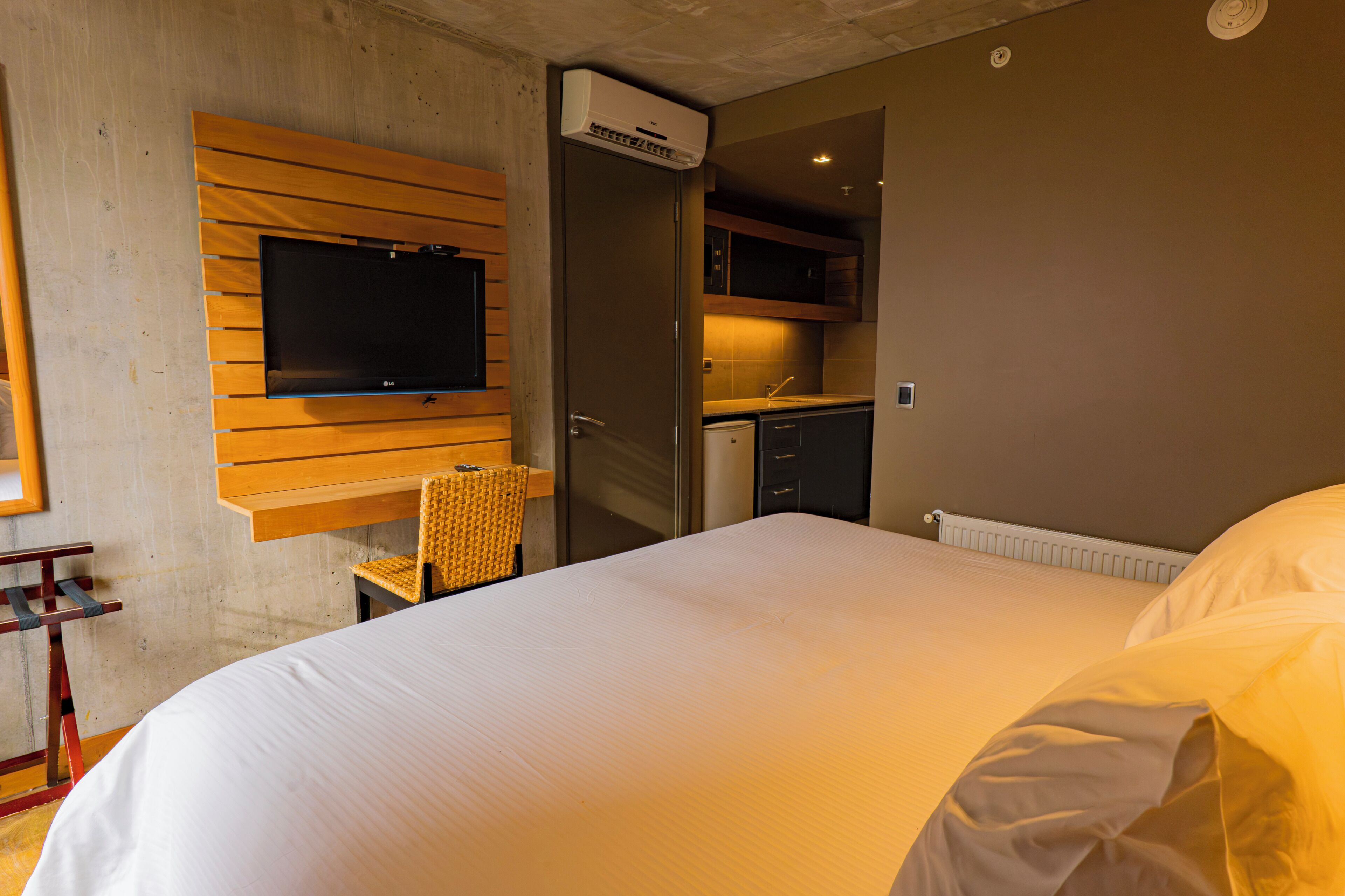 Una habitación de hotel con una cama, televisor montado en un panel de madera, kitchenette y una silla.