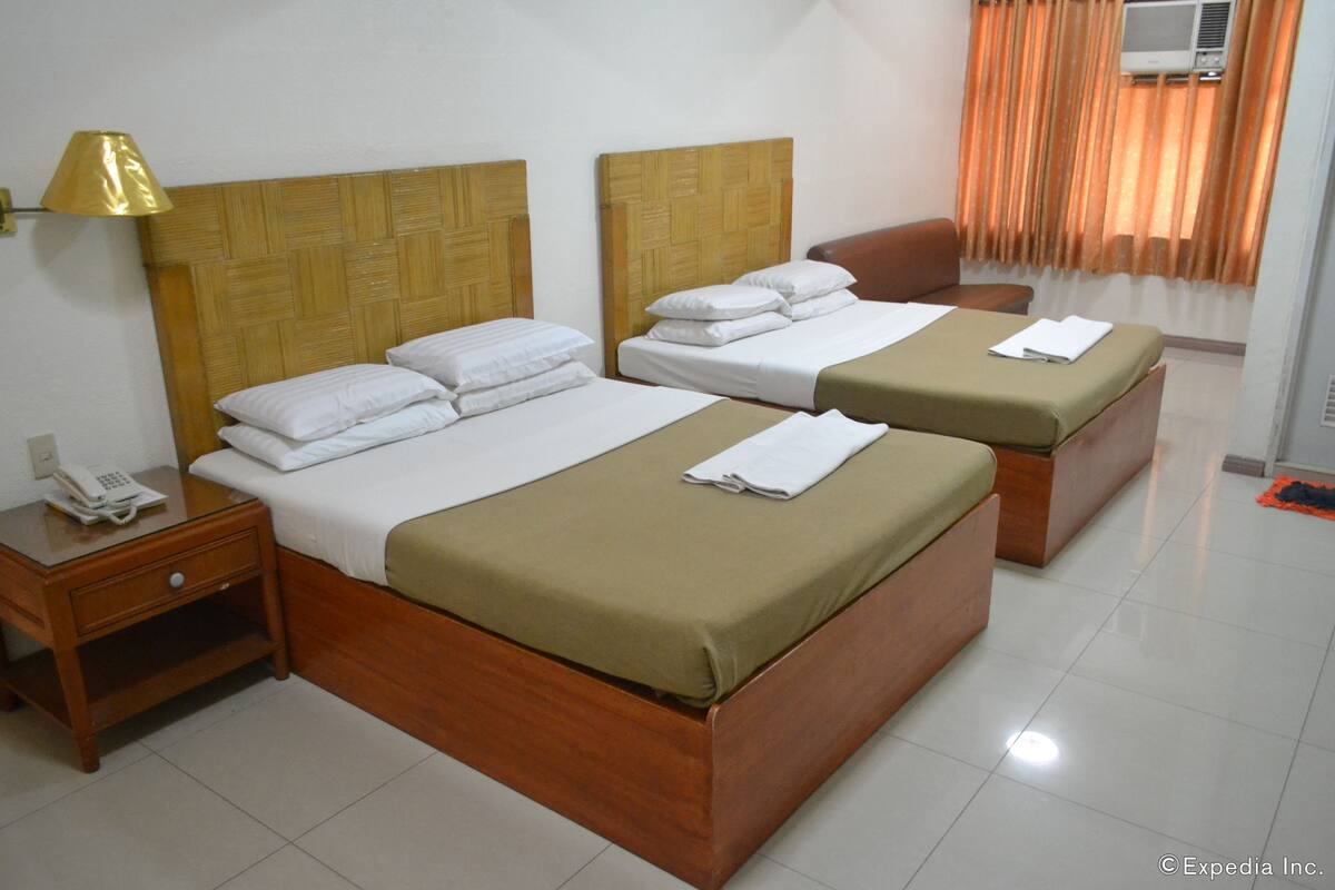 A hotel room with two beds, a wooden headboard, a bedside table with a lamp, and a small table with a telephone.