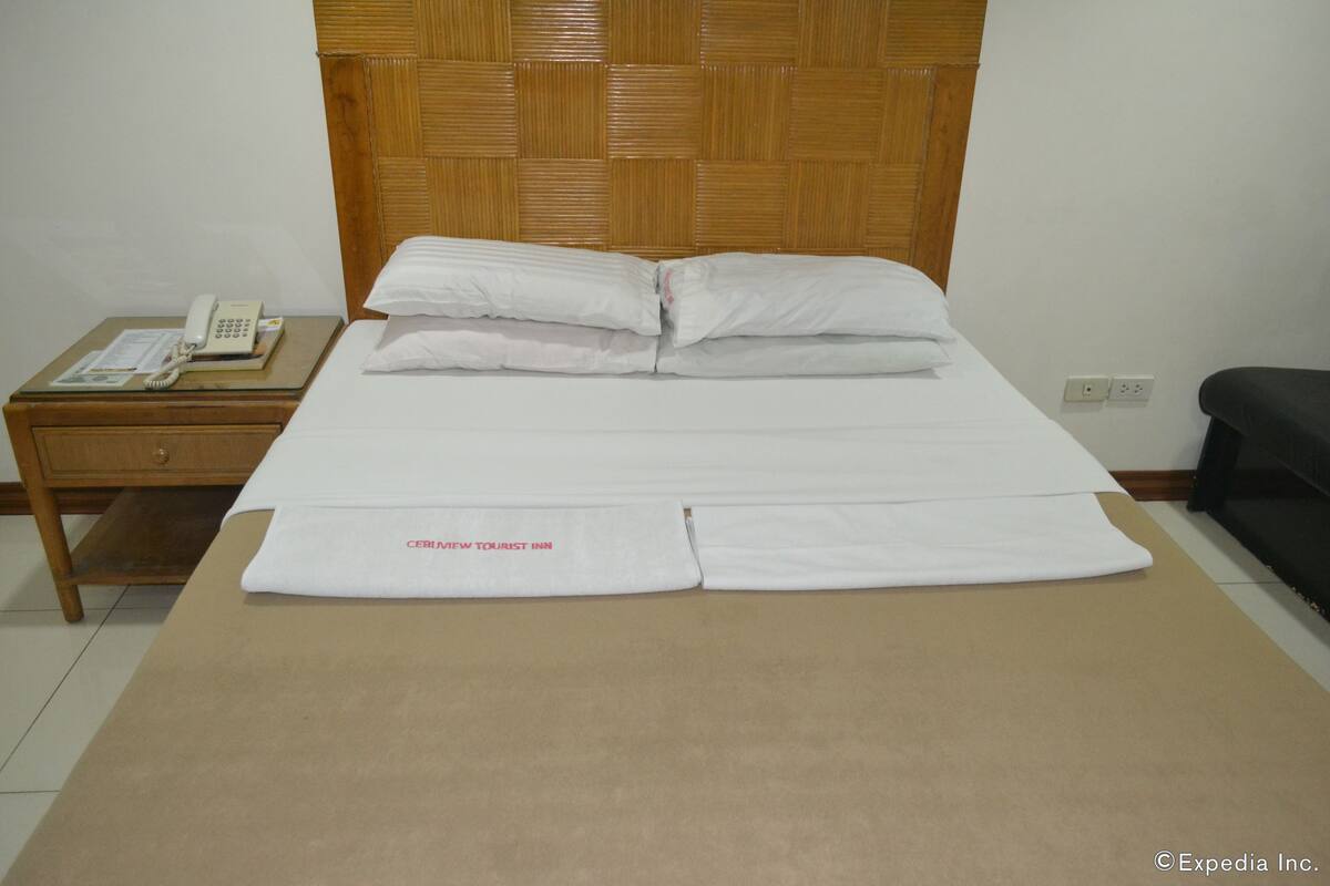 A neatly made bed with white linens and a folded towel on the bed. There is a wooden headboard, a bedside table with a telephone, and a power outlet on the wall.