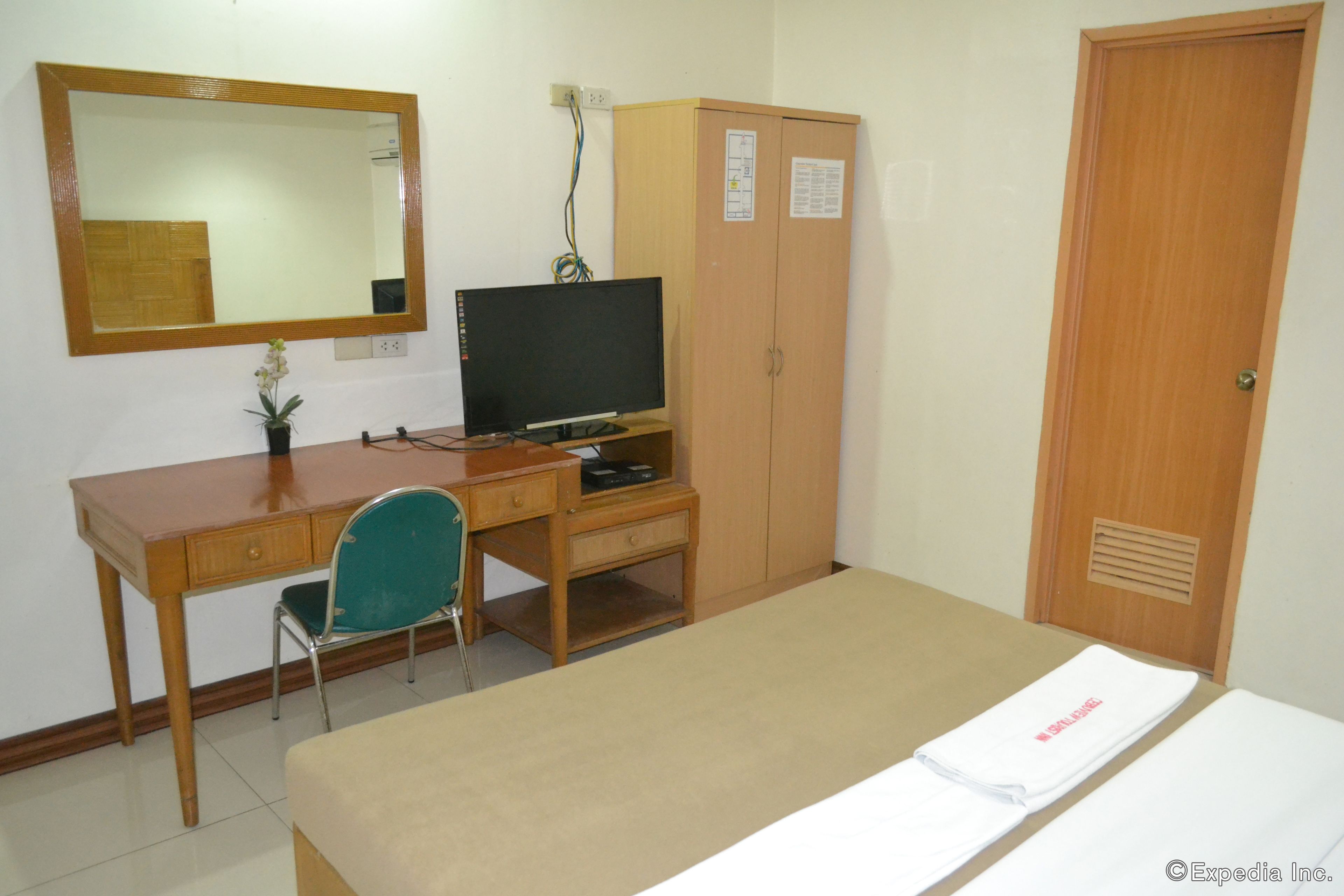 Una habitación con escritorio de madera, una silla verde, televisor, armario y cama con manta blanca.
