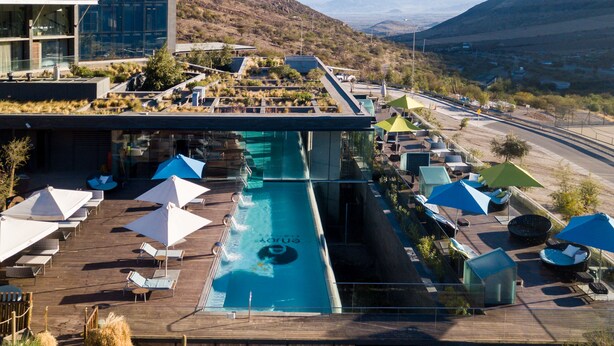 Ein Dachpool mit Liegestühlen und Sonnenschirmen, umgeben von einem modernen Gebäude mit Glasfassade.