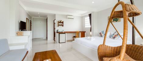 A modern hotel room with a large bed, a hanging chair, a small kitchenette, and a seating area with a sofa and table.