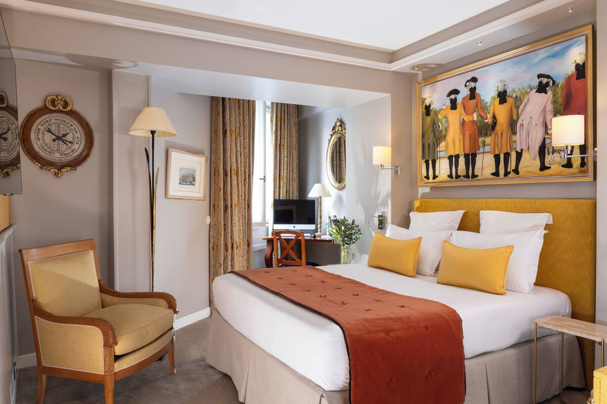 A hotel room with a large bed, a wooden chair, a desk with a lamp, a painting on the wall, and a clock.