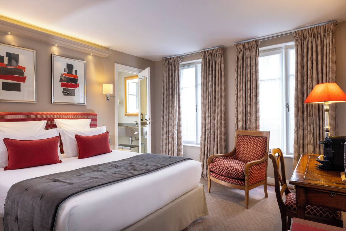 A hotel room with a large bed, red pillows, a wooden chair, a desk with a lamp, and a window with curtains.