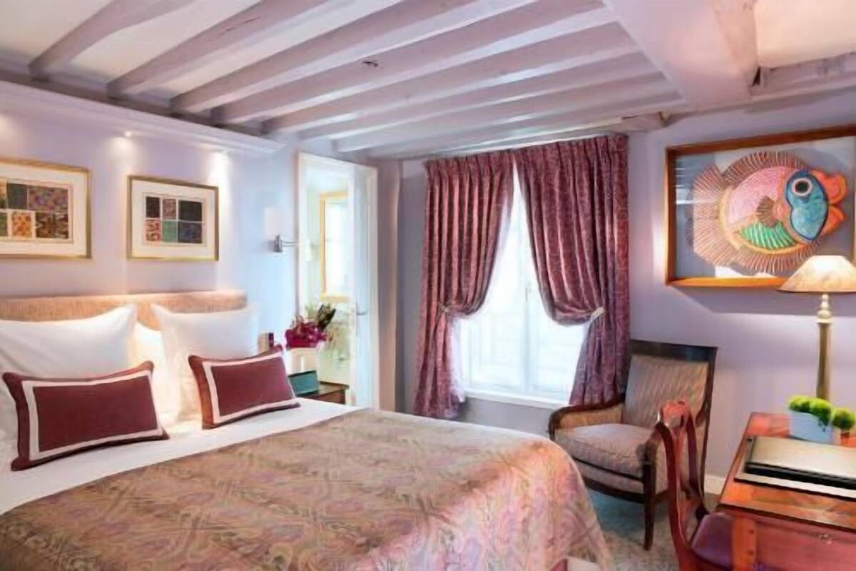 A hotel room with a bed, a chair, a desk, a window with curtains, and artwork on the walls.