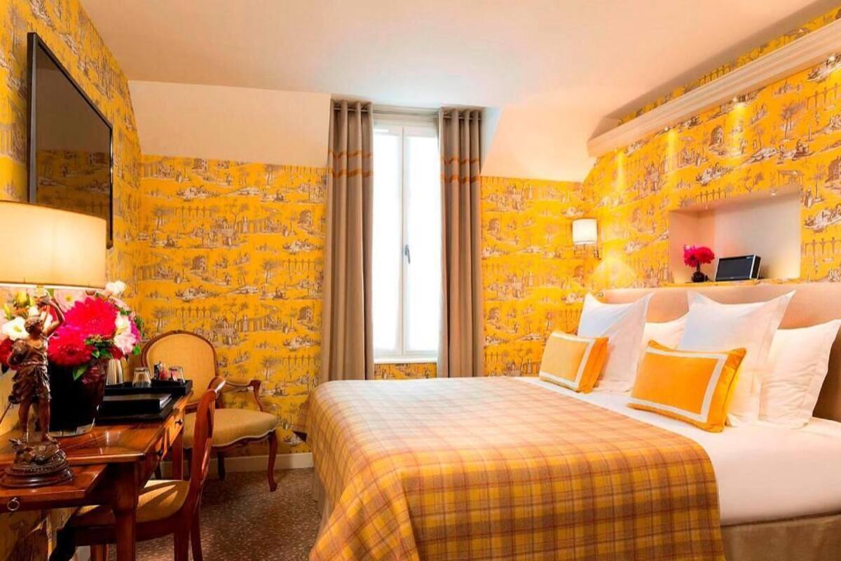 A hotel room with a bed, a desk with a chair, a vase of flowers, and a window with curtains.