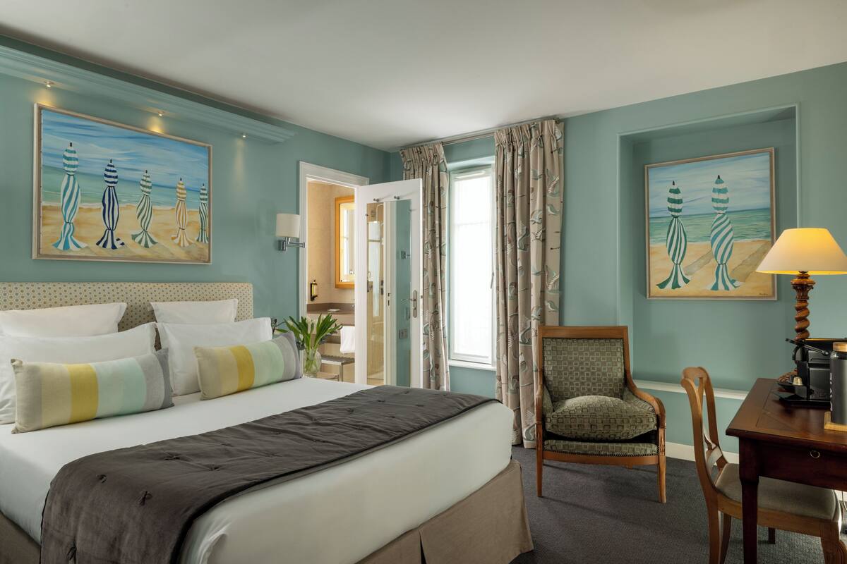 A hotel room with a bed, a chair, a desk, and two paintings on the walls.