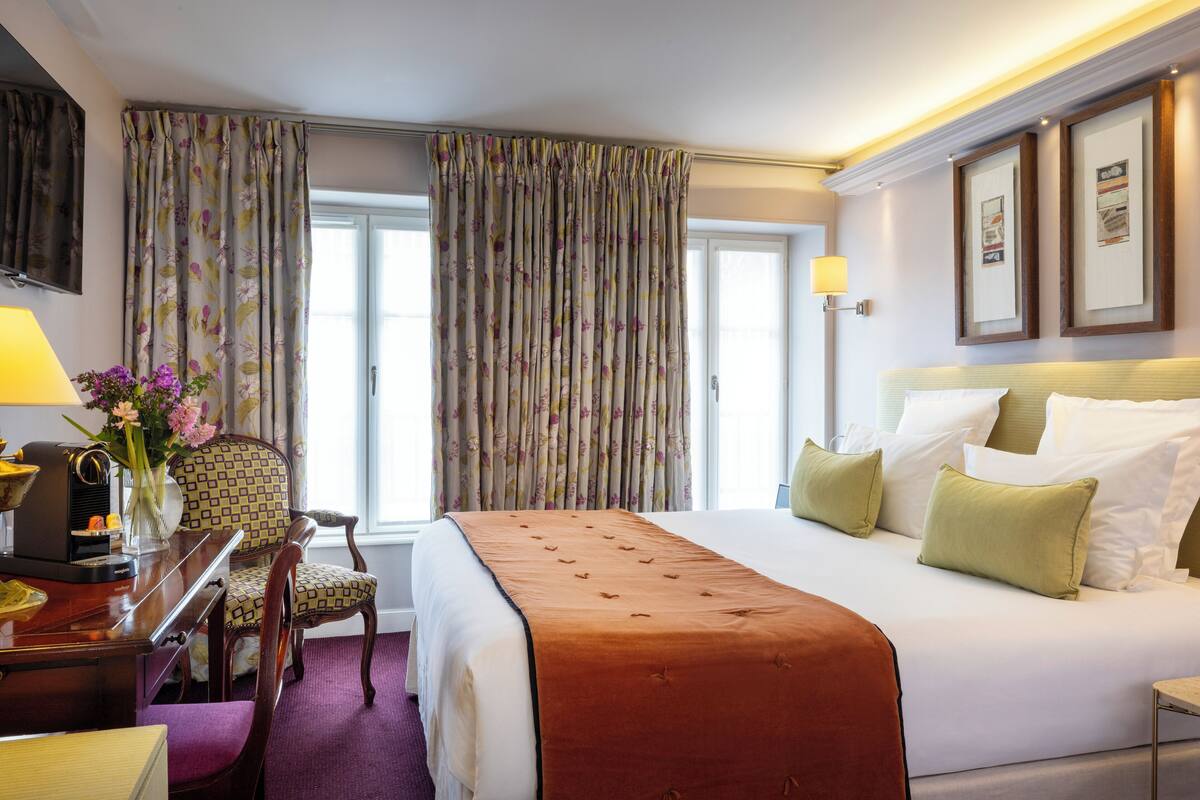 A hotel room with a large bed, a desk with a lamp, a chair, and a window with curtains.