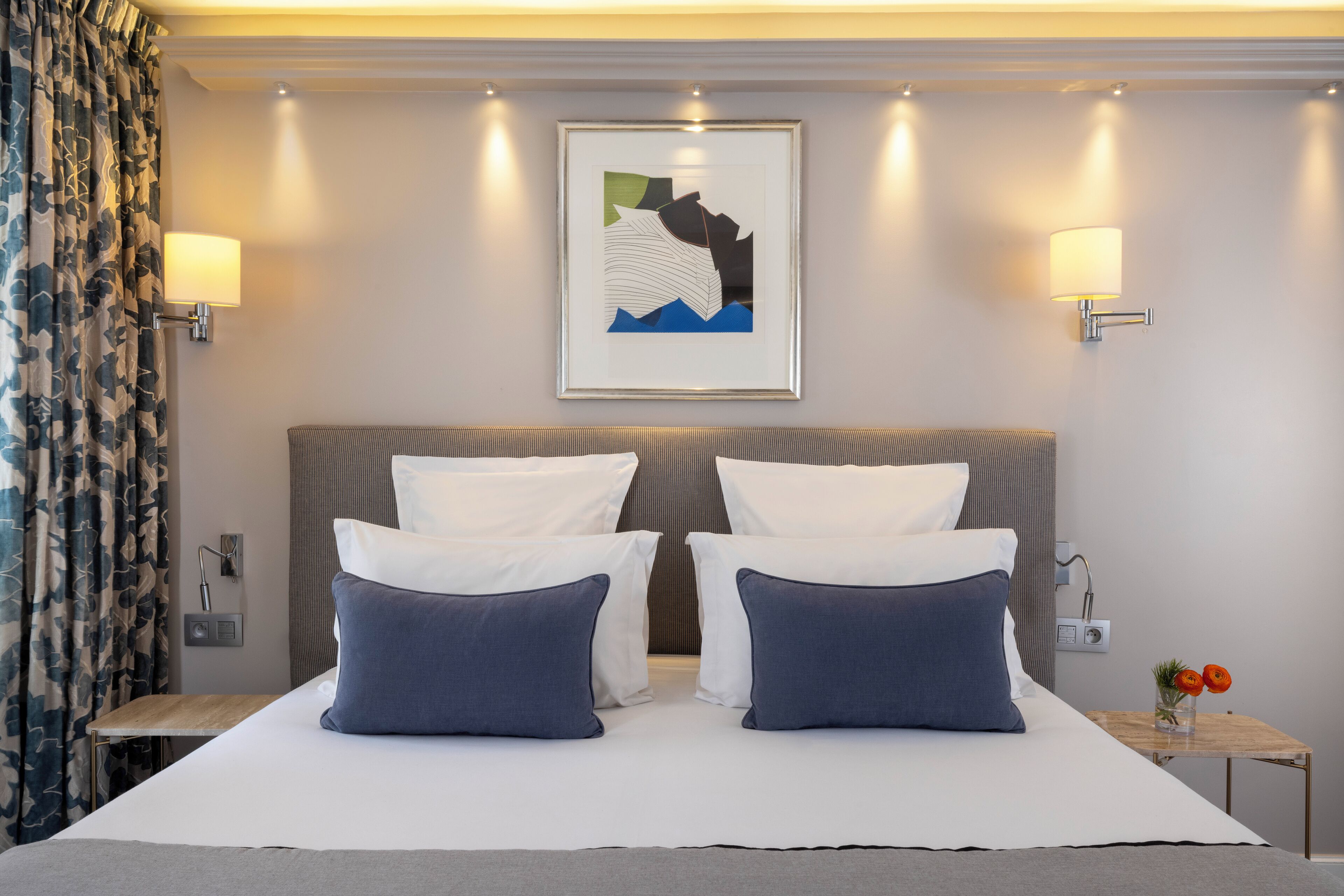 Een hotelkamer met een groot bed, twee bedlampen, een kunstwerk in een lijst aan de muur, en een tafeltje met een bloemenvaas.