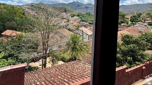 A view from a window overlooking a town with red-roofed buildings, greenery, and mountains in the distance. - Pousada Casa dos Contos (Ouro Preto)