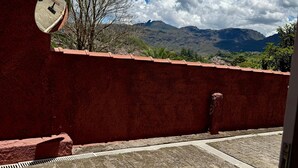 A courtyard with a red wall, a tiled floor, and a view of mountains and trees under a blue sky with clouds. - Pousada Casa dos Contos (Ouro Preto)