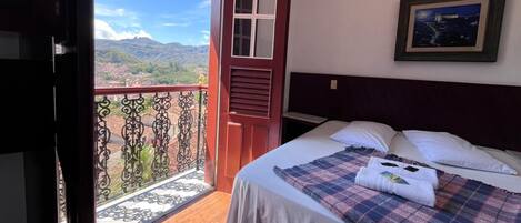A bedroom with a bed, a nightstand, a framed picture on the wall, and a balcony with a view of the mountains.