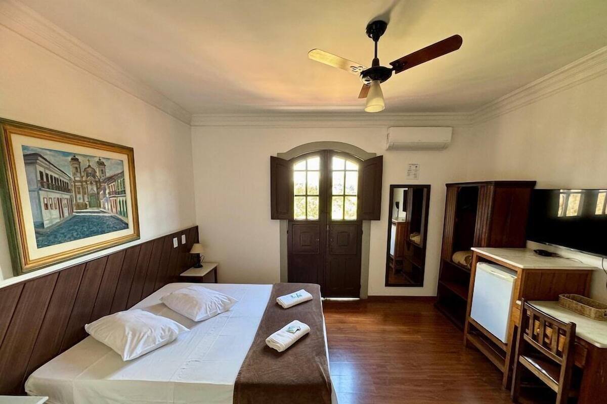 Habitación de hotel con cama, televisor, ventilador de techo, pintura, ventana y puerta.