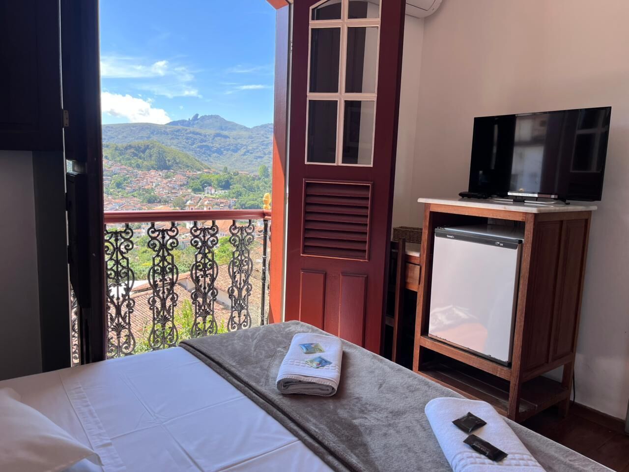 A bedroom with a bed, a TV, a mini-fridge, and a balcony with a view of the mountains.