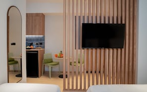 A modern hotel room with a wooden partition, a flat-screen TV, a kitchenette, and two green chairs.