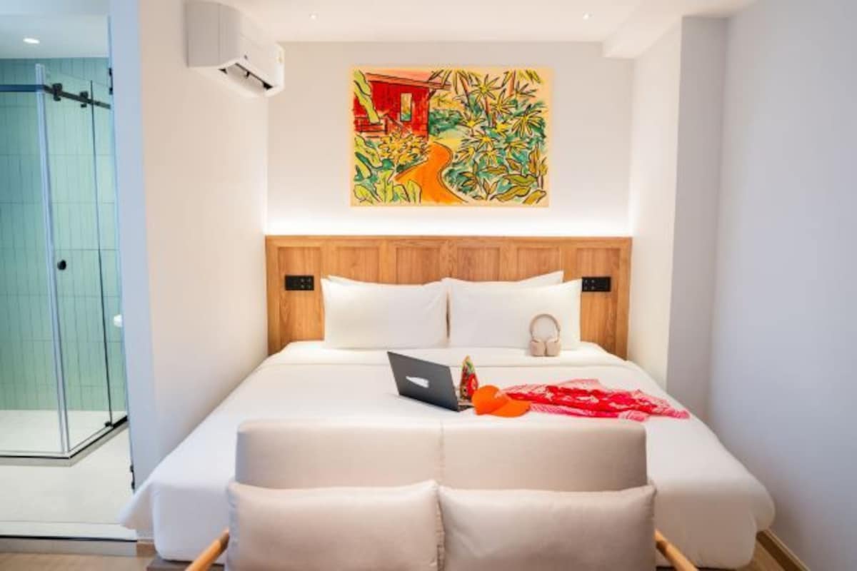 Una habitación de hotel moderna con una cama grande, un ordenador portátil sobre la cama, un cuadro en la pared y una zona de ducha.