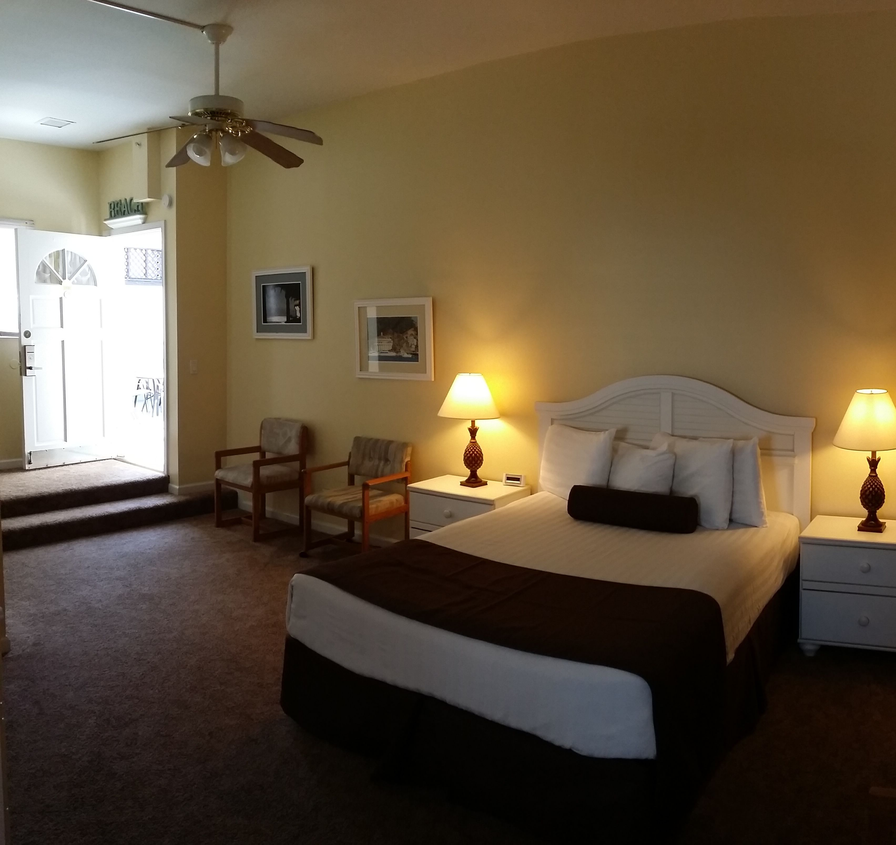 A hotel room with a bed, two bedside tables with lamps, a chair, a ceiling fan, and a door leading to a balcony.