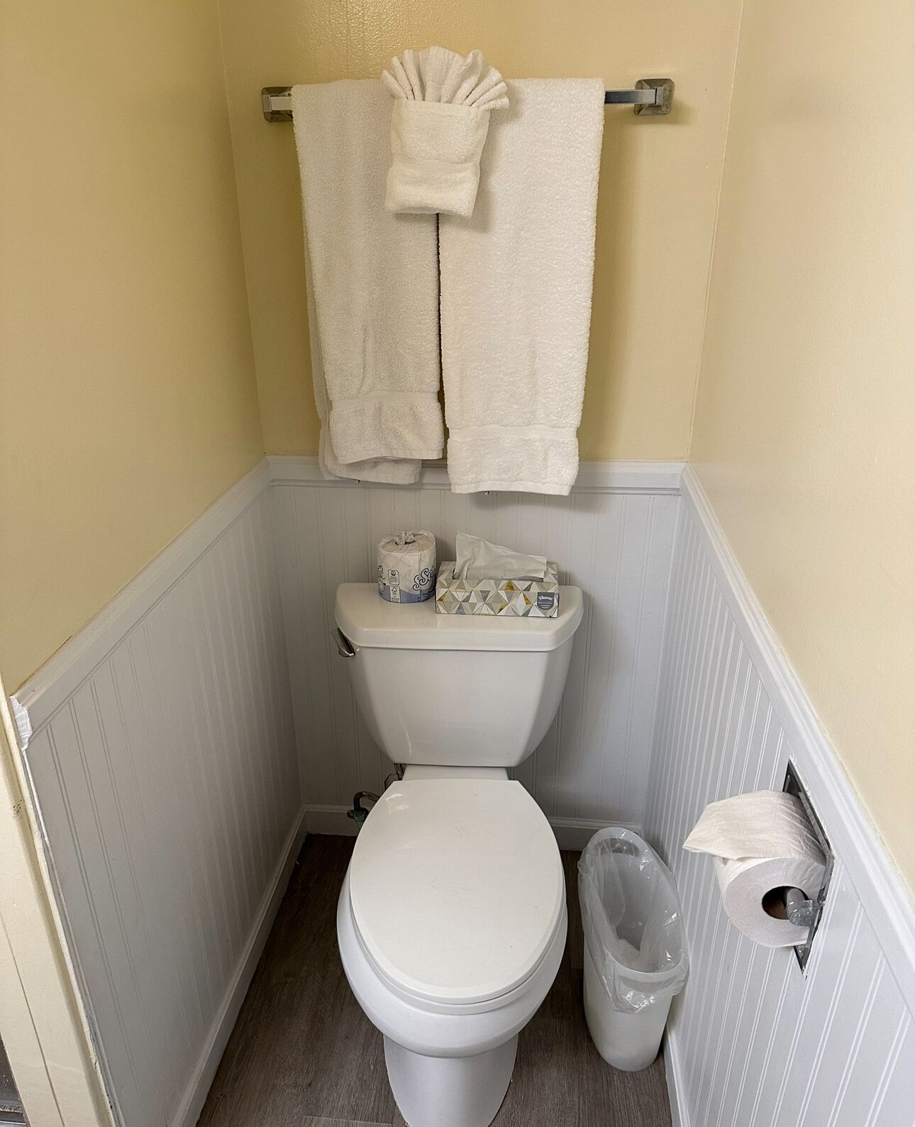 Un baño pequeño con un inodoro, dos toallas blancas colgadas en un perchero metálico, un rollo de papel higiénico y un cubo de basura.