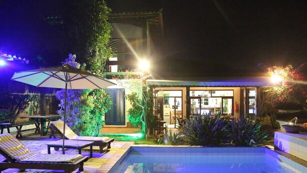 Zona de piscina con sillas de descanso y una mesa, rodeada de plantas y un edificio con entrada visible.