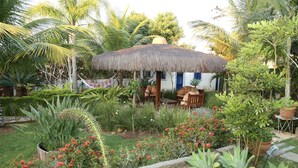 Una cabaña con techo de paja y un jardín delantero, rodeada de plantas tropicales y una hamaca.