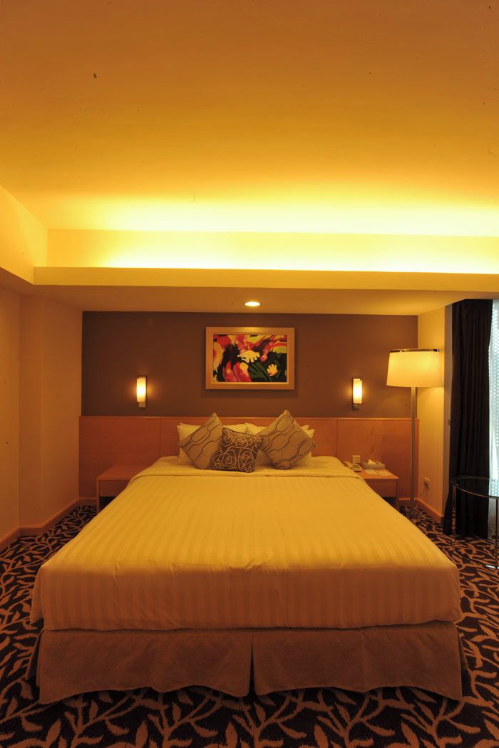 A hotel room with a large bed, a headboard, a painting on the wall, a bedside lamp, and a patterned carpet.