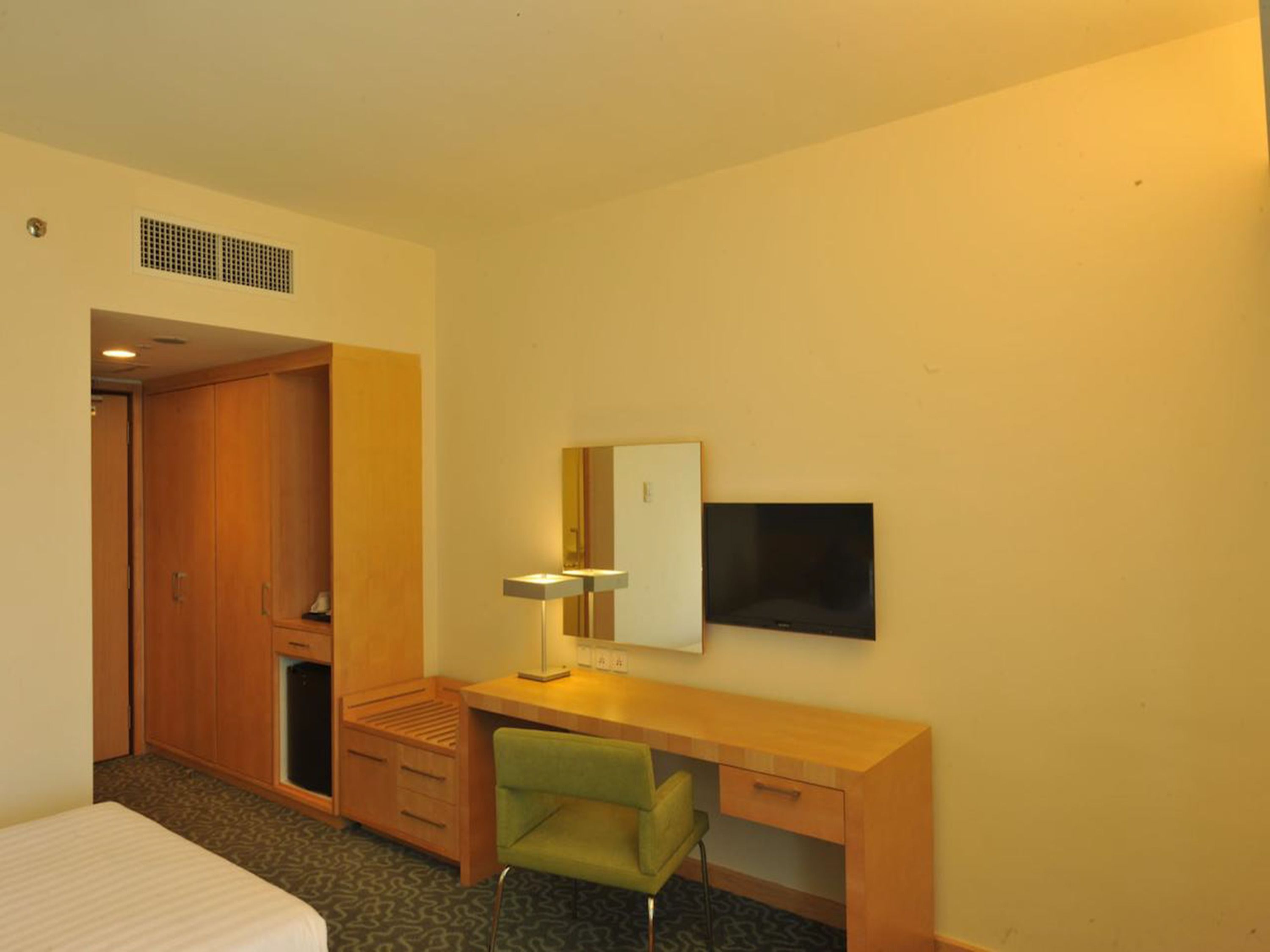 A hotel room with a wooden desk, a green chair, a flat-screen TV, a mirror, and a wardrobe.