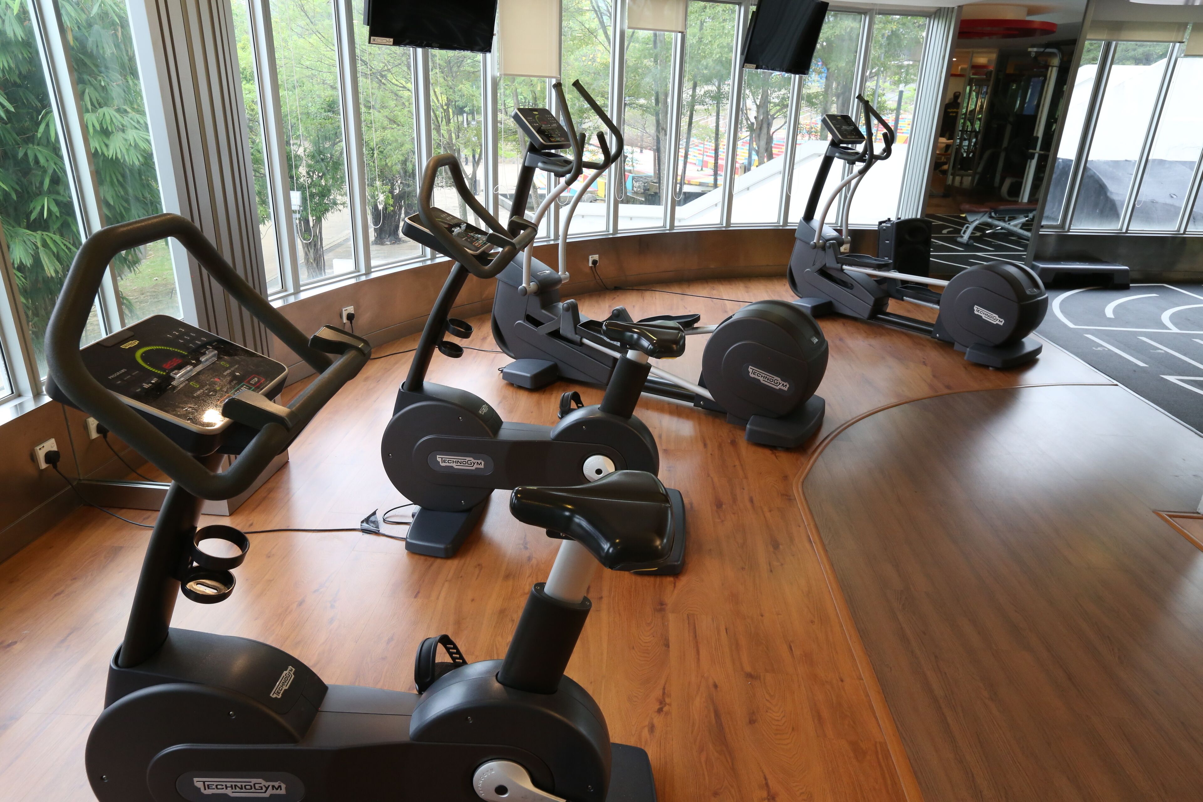 A gym with cardio machines, large windows, and a wooden floor.