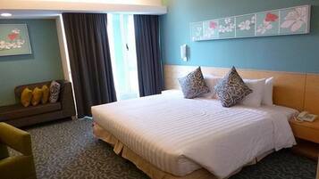 Ein Hotelzimmer mit einem großen Bett, einer Couch und einem Sessel. Es gibt ein Fenster mit Vorhängen, eine Wand mit Blumenbild und ein Telefon auf dem Nachttisch.