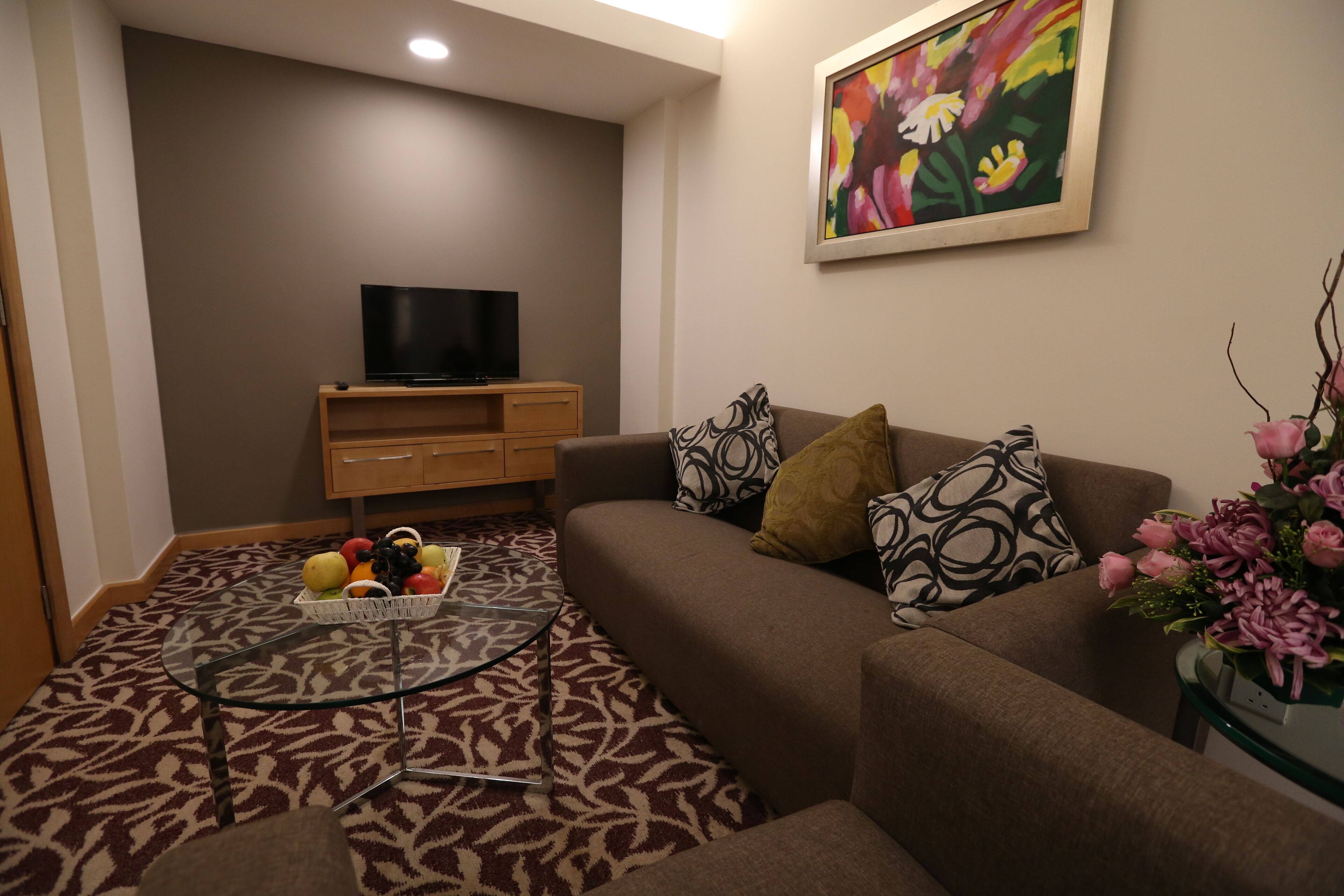 Una sala de estar con un sofá, una mesa de centro de vidrio, una cesta de frutas, un televisor sobre un mueble de madera y un cuadro en la pared.