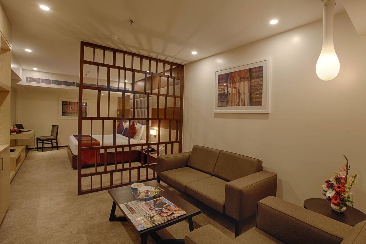 A hotel room with a bed, a sofa, a coffee table with a magazine, a desk with a chair, and a decorative partition.