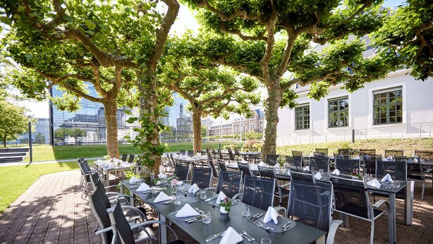 An outdoor dining area with tables and chairs, surrounded by trees and a building in the background. - Kasino Hotel (Leverkusen)